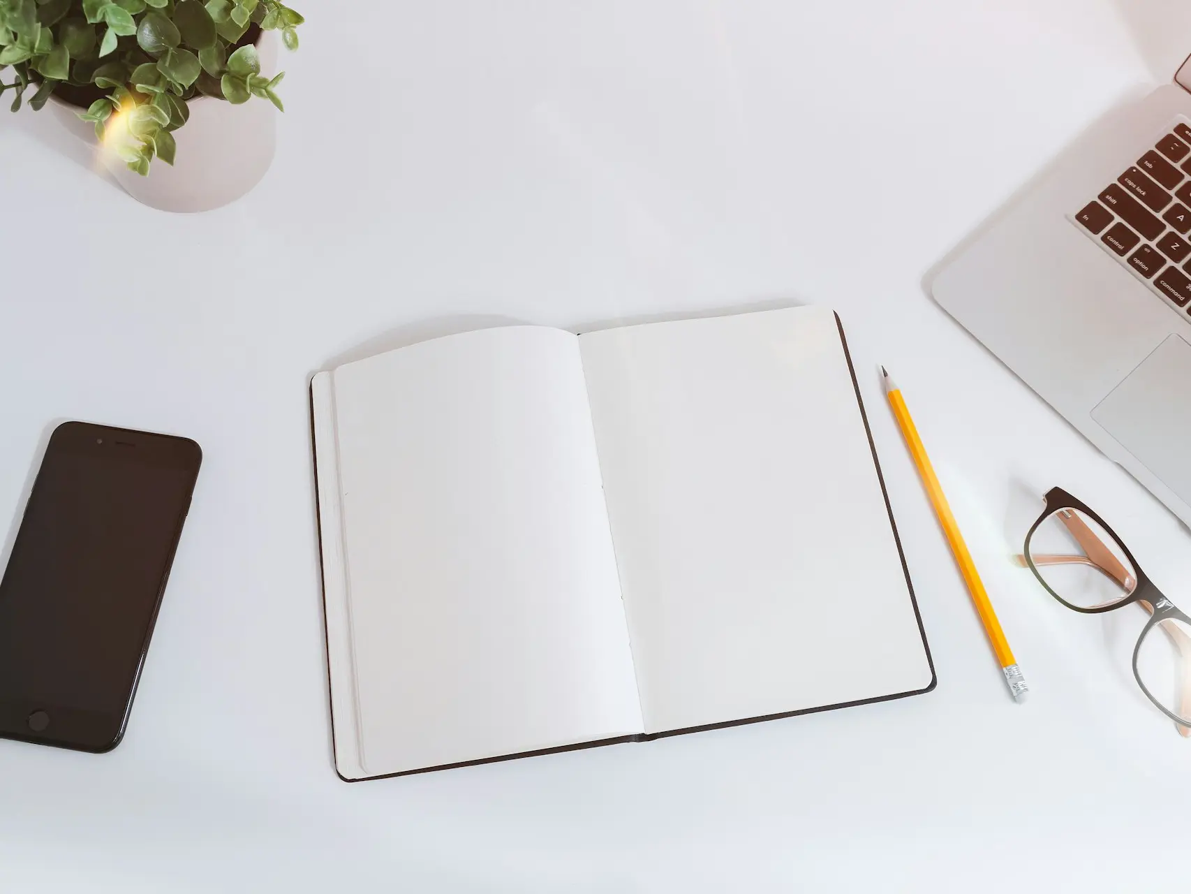 An open empty notebook on a white desk next to an iPhone and a MacBook